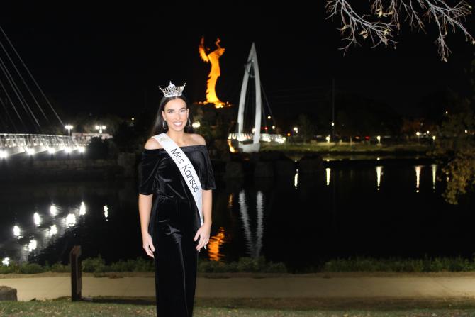 Miss Kansas poses across the river from the Keeper at Night which is illuminated in a nighttime scene