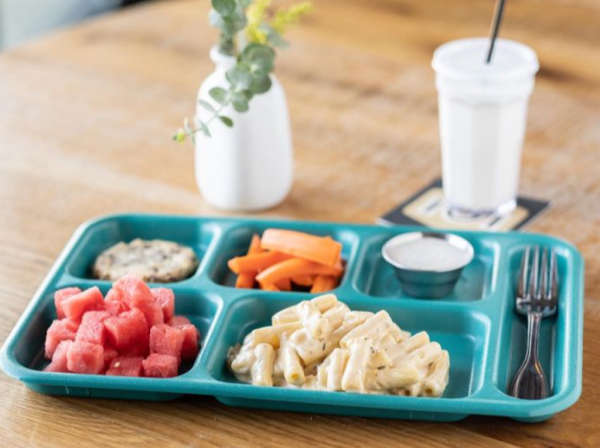 A kid's meal from 101 Beer Kitchen, served on a teal vintage lunch tray. The meal includes creamy macaroni and cheese, fresh watermelon chunks, carrot sticks, a cookie, and a small cup of ranch dressing. A white milkshake with a black straw is placed on a wooden table next to a small white vase with green foliage.