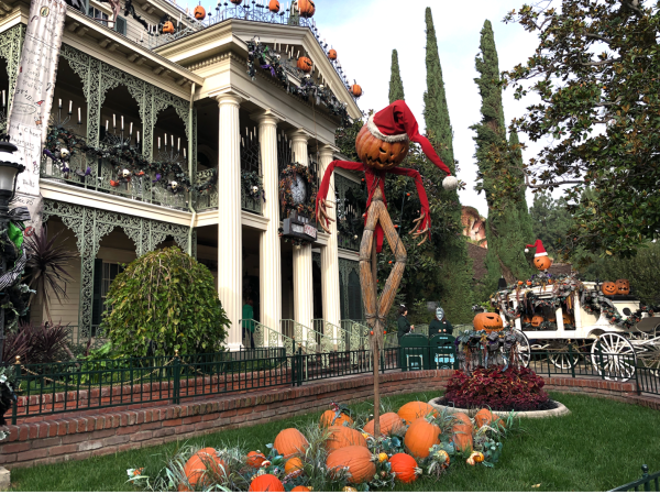 Image of the front of Haunted Mansion Holiday at Disneyland Resort. A large, white mansion can be seen in the background. In the foreground, a scarecrow with a pumpkin head can be seen.