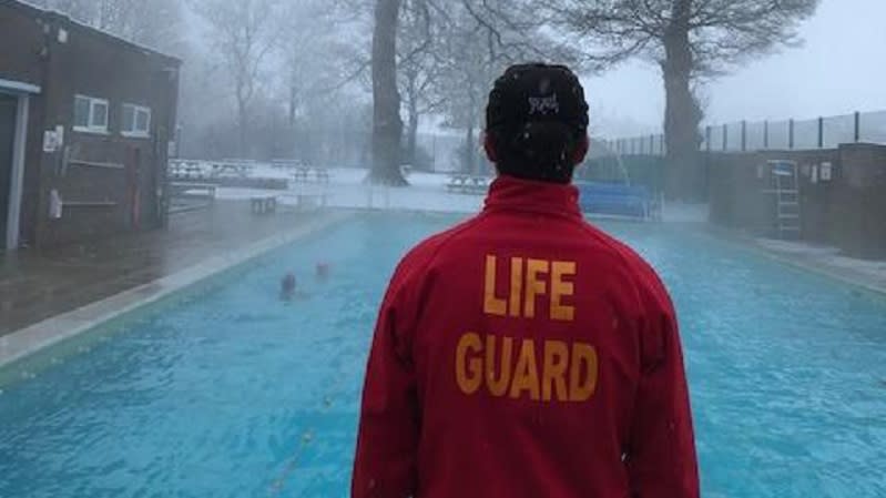 A lifeguard stands, with his back to the camera, looking at the swimming pool with steam rising into the cold air, surrounded by snow dusted ground