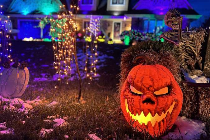 A Halloween Jack-o-Lantern is lit up, with a home that's been decorated for Halloween in the background. Also in the background are more halloween lights, skeletons, and other ghoulish delights.