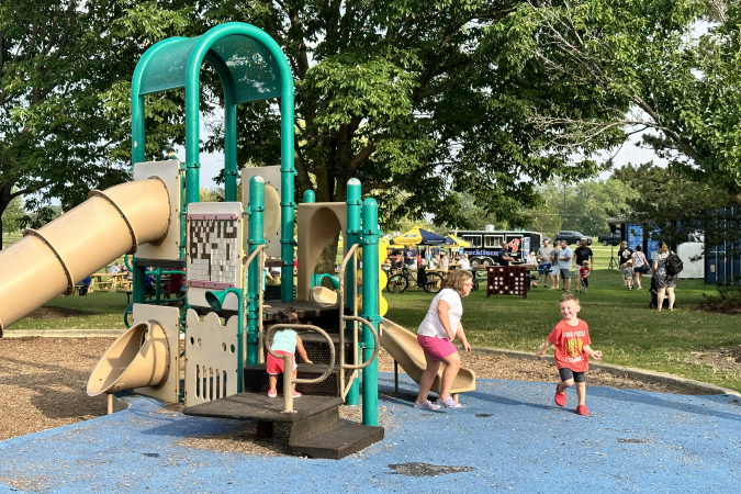 Children play on the playground near the Lake Andrea Beer Garden, with the beer garden picnic tables visible in the background.