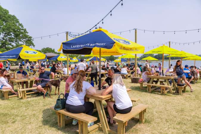 Patrons sitting at picnic tables at Lake Andrea Beer Garden with sunny skies and blue water in the background