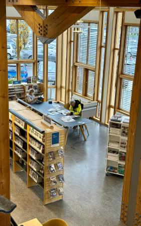 To personer sitter ved et bord og leser aviser i et lyst bibliotek med store vinduer og bokhyller.