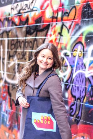 A woman stands in Graffiti Alley in Ann Arbor holding a reusable bag that features the Destination Ann Arbor logo.
