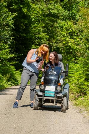 A lady using a mobility scooter in the forest, whilst her friend shows her a photo on the camera