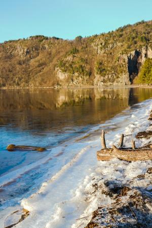 Islagt strand ved et stille vann med fjell og skog i bakgrunnen.