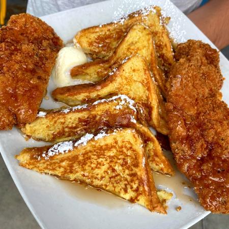 Image of five slices of french toast with two slices of fried chicken on the left and right sides. Food is topped with maple syrup, powdered sugar and butter.