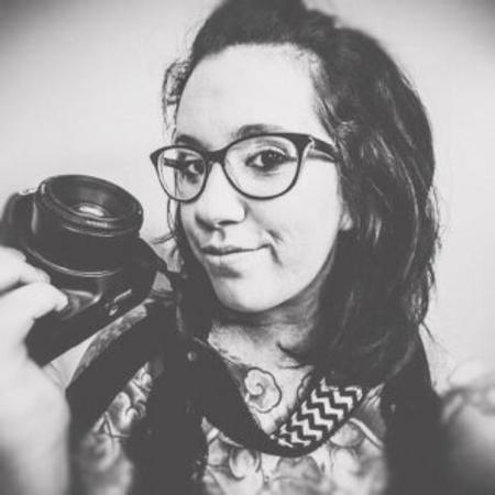 A photo of a woman, Nicole Caracia, in black and white. She has dark hair, wears glasses and is holding a camera