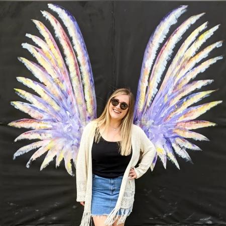 A smiling woman in sunglasses poses in front of a colorful painted mural of angel wings on a black backdrop in Fayetteville, NC. The wings are lavender with accents of yellow, pink, and white, creating a vibrant, photo-worthy moment.