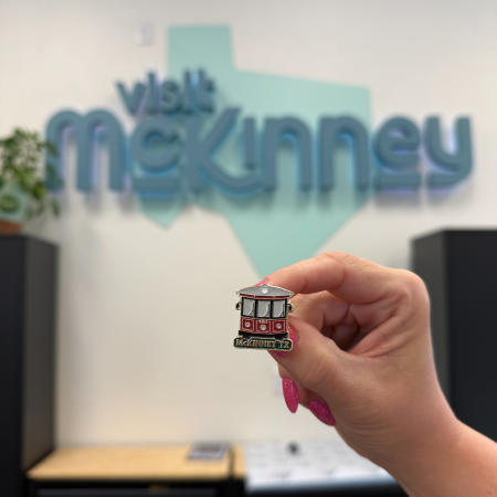 Red and green trolley enamel pin in front of Visit McKinney store sign