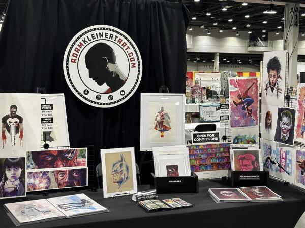Artist booth for Southern Indiana artist Adam Kleinert displaying original artwork, prints, and commissions at a convention, featuring colorful paintings of comic book, movie, and pop culture characters.