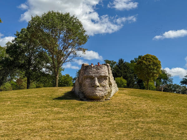 Stone sculpture of a human face built into a grassy hillside, surrounded by trees under a clear blue sky with scattered clouds. Visitors can be seen at the top of the sculpture, adding a sense of scale to this unique outdoor artwork.