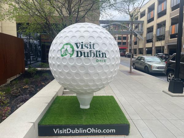 Oversized golf ball sculpture with “Visit Dublin Ohio” branding displayed on a sidewalk in Downtown Dublin near parked cars and buildings.