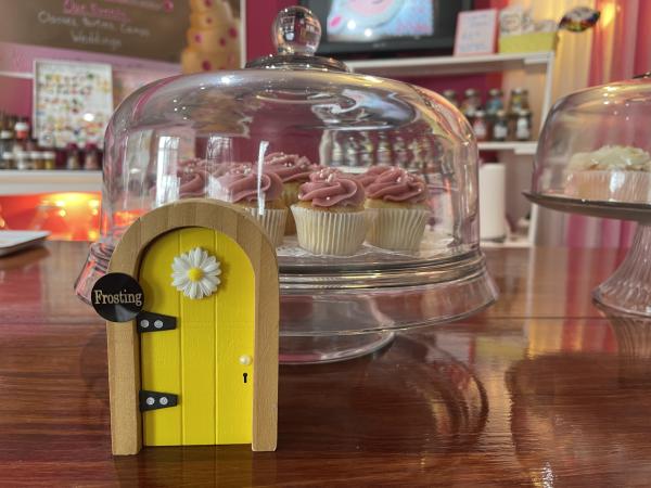 Yellow fairy door in front of cupcakes in a glass case at Our CupCakery