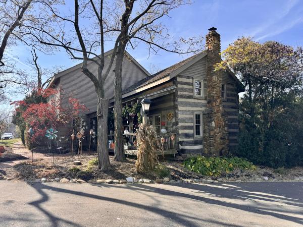 The exterior of The Morgan House in Dublin, Ohio, highlighting its unique log cabin construction and stone chimney. The building is surrounded by colorful fall foliage and decorative items, creating a cozy and inviting atmosphere.