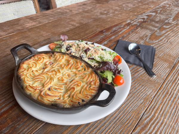 A plate featuring a shepherd's pie in a cast-iron dish with swirled mashed potato topping, served alongside a mixed green salad with cherry tomatoes, almond slivers, and shredded cheese on a rustic wooden table.