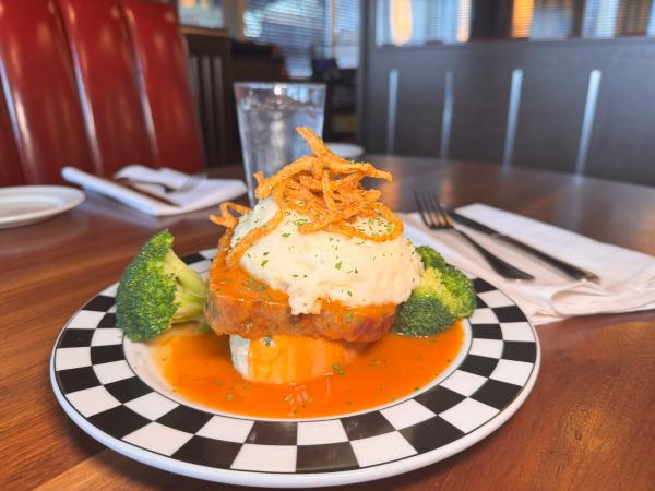 A plated meatloaf dish from Cap City, featuring a slice of meatloaf topped with mashed potatoes, crispy fried onions, and garnished with parsley. The dish is served with broccoli florets and surrounded by a vibrant orange sauce on a black-and-white checkered plate, set on a wooden table in a diner-style setting.