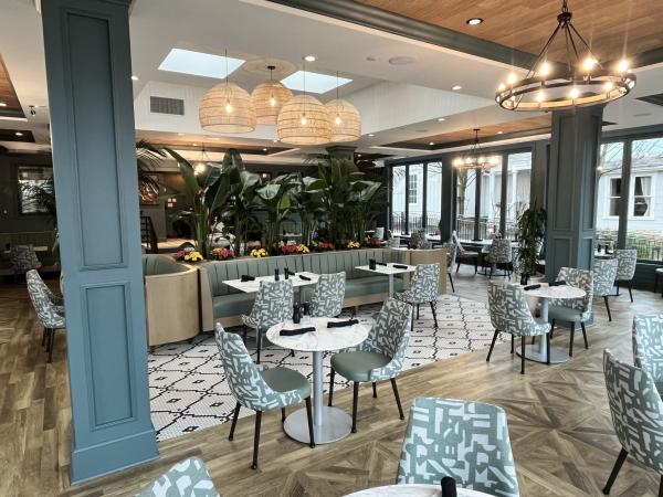 A stylish restaurant dining area with soft green and white patterned chairs, round marble tables, and cozy booth seating. Large windows let in natural light, while lush plants, pendant lights, and chandeliers enhance the elegant ambiance.