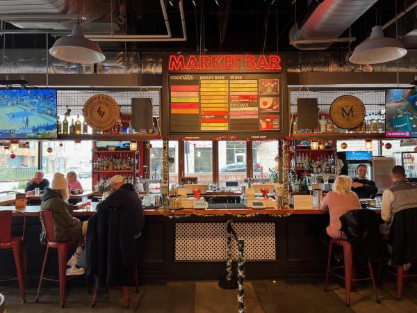 Market Bar Interior: A vibrant bar setup with a glowing "MARKET BAR" sign overhead. Patrons are seated at the bar enjoying drinks, surrounded by shelves stocked with bottles and a colorful drink menu board displayed prominently. Sports games play on mounted TVs, completing the lively atmosphere.