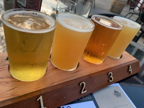 Beer Flight (North High Brewing): A wooden tray labeled with numbers 1 to 4 holds four small glasses of beer, each showcasing a different hue, from pale gold to rich amber. The beers appear freshly poured, with bubbles rising to the surface. A relaxed outdoor setting with a faint reflection of sunlight and patio furniture in the background enhances the casual atmosphere.