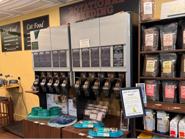 Section of Pet Wants store featuring bulk pet food dispensers labeled for rotational feeding. A sign explains the benefits of small-batch, clean-ingredient food. Packaged pet food bags are neatly displayed on nearby shelves.
