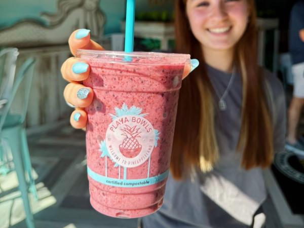 A cheerful young woman holding a Playa Bowls smoothie in a clear cup, topped with a blue straw. The background showcases the colorful tropical-themed interior of the shop.