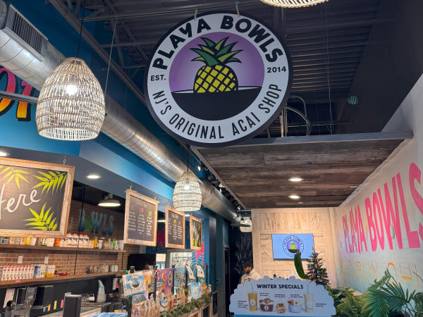 The interior of a Playa Bowls shop, featuring a rustic tropical decor with wicker light fixtures, colorful signage, and a prominent logo that says "Playa Bowls, NJ's Original Acai Shop."