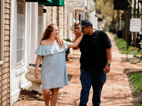 A man and woman walk down a brick sidewalk in Historic Dublin, Ohio. The man kisses the woman's hand as they stroll past shops and trees.