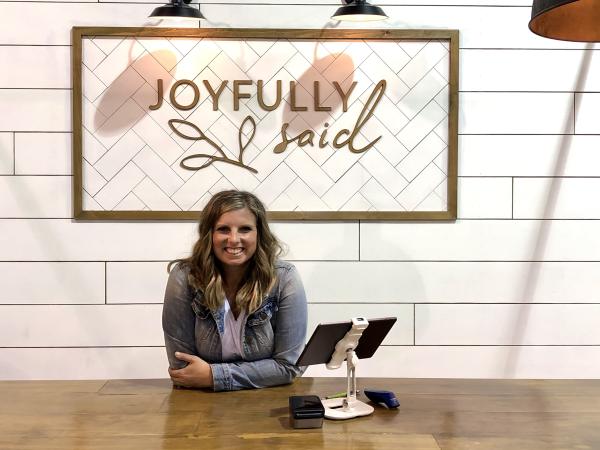 Chelsea Gorsuch sits at a desk with a sign on the wall behind her that reads "Joyfully Said"