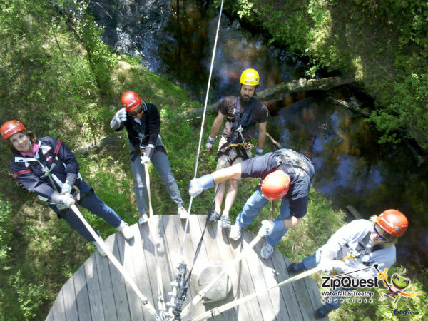 Group of people wearing helmets and safety harnesses standing on a zipline platform at ZipQuest, surrounded by trees and water below.
