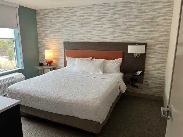 Modern hotel room with a neatly made bed, white bedding, wood headboard, bedside lamps, textured accent wall, and a window providing natural light.