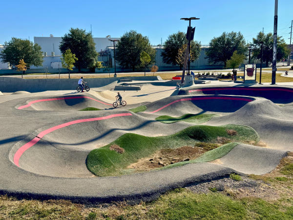 A wavy asphalt pump track sits in the foreground while the children bike through the skatepark at Riverfront in Fort Smith, Arkansas.