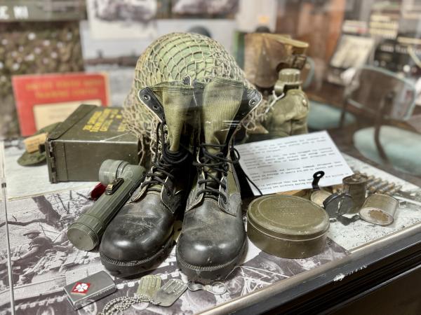 Black leather boots sit behind a flip lighter and worn dog tags, an ammo crate, helmet, and canteen are also curated within this display at the Chaffee Barbershop & Military Museum.