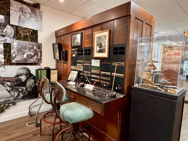 A historic military switchboard console is inlayed into a large wooden frame and includes two operator chairs.