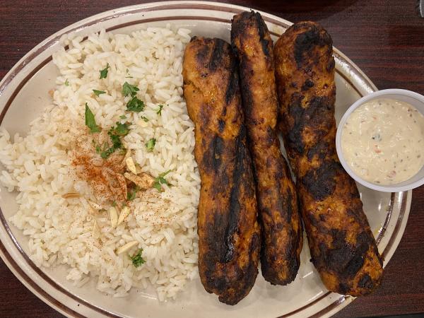 An overhead view of a plate from Al Shish Palace in Huntsville, Alabama, featuring three grilled beef kofta kebabs served with seasoned rice topped with sliced almonds, parsley, and paprika, alongside a cup of creamy garlic sauce.