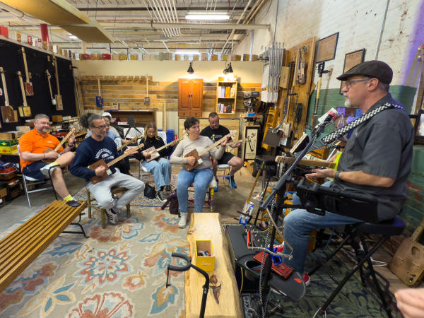 Participants gather inside the Cigar Box Guitar Store at Lowe Mill ARTS & Entertainment for a group lesson on playing cigar box guitars. An instructor leads the class while students sit in a circle holding handcrafted instruments, surrounded by workshop tools, guitar displays, and shelves filled with cigar box guitar materials.