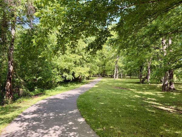 A paved walking trail winds through the Indian Creek Greenway in the Village of Providence in Huntsville, Alabama, surrounded by large trees, shaded grassy areas, and lush greenery on a sunny day.