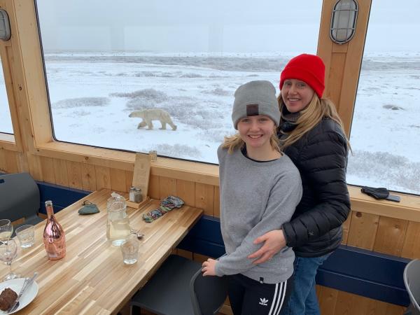 A blonde woman and her daughter smile from inside a window with a polar bear visible outside.