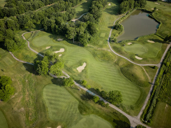 An aerial view of a golf course with several greens, fairways, sand bunkers, cart paths, trees, and a large pond.