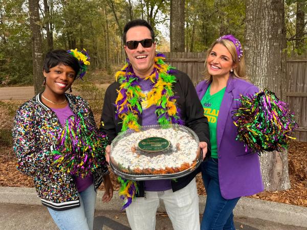 Podcast hosts Zondra and Katie stand on either side of baker Zach Zornes of Randazzo's Camellia City Bakery. He holds a king cake toward the camera.