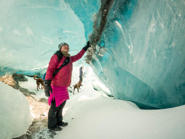 a woman touches the frozen walls of a glacier ice cave. Dogs in background.