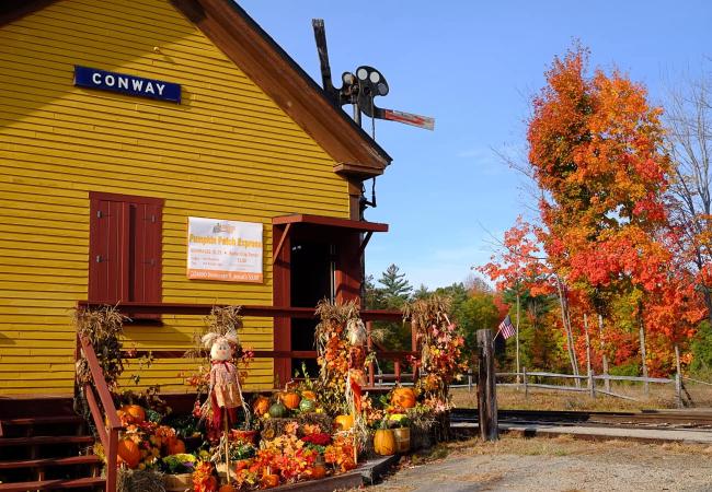 Fall decorations outside a yellow train station with the word "Conway" in white on a blue sign