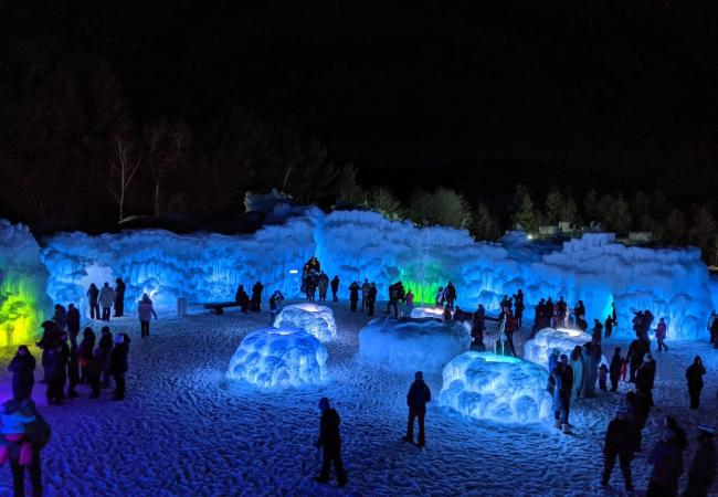 A nighttime scene of a crowd of people mingling inside a large space surrounded by ice walls lit by LED lights