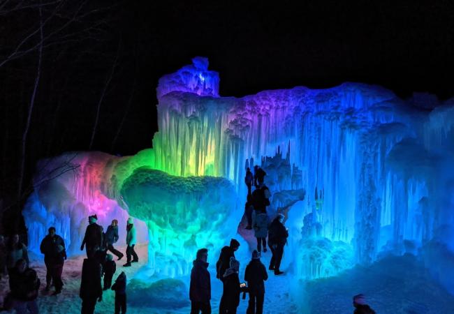 A group of people emerge from a tunnel carved in ice and colored with red, blue and yellow lights Ice Castles