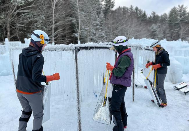 three people in winter clothes and helmets gather icicles from a metal frame to use in building New Hampshire's Ice Castles