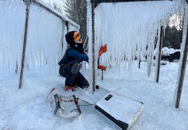 A person in winter clothes and gloves pulls icicles that are hanging from a metal pipe