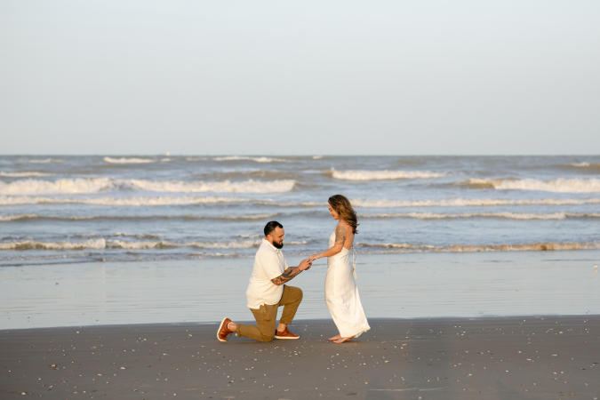 A man kneels on the beach to propose to a woman in a white dress at sunset, with gentle waves and a calm ocean in the background on South Padre Island.