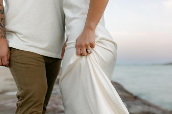 Close-up of a couple standing on the beach, highlighting the woman’s hand gently holding her white dress and displaying a large engagement ring, with ocean waves and a pastel sky in the background.
