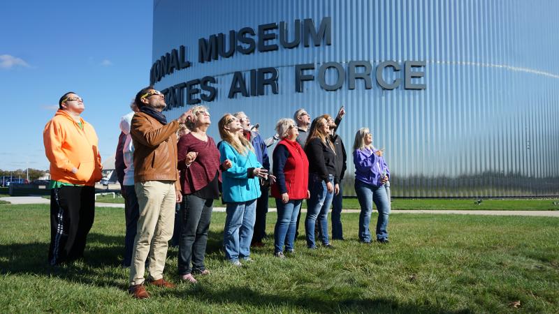 A diverse group of individuals, all wearing solar eclipse glasses and standing in front of the National Museum of the U.S. Air Force, looking skyward to the right, as if viewing a solar eclipse.
